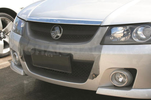 Close-up of a silver car with a Mean Mother 4x4 Radiator Bug Protector (90cm x 60cm) installed on the front, covering the grille. The Holden badge, blacked-out license plate, headlights, and fog lights are visible; another car’s wheel appears at left.