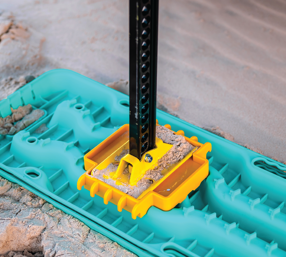 An Exitrax Ultimate 1150 Recovery Board in Aqua Marine is used under a car jack on sandy terrain. The secure yellow base of the jack holds firmly on the board, preventing it from sinking into the sand.