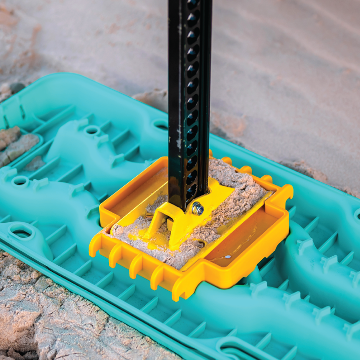 Close-up of a construction jack footplate with a sandy base. The footplate consists of a yellow metal bracket holding a black, perforated jack pole, mounted onto a teal plastic platform. Equipped with traction enhancement comparable to the Exitrax Recovery Board Ultimate 1150 - Blood Orange from Exitrax, the setup suggests an outdoor construction setting.