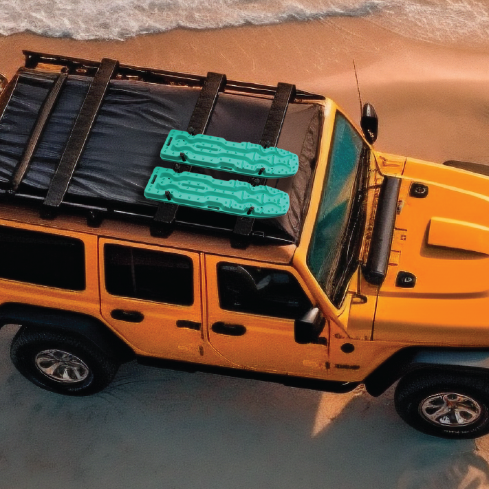 An orange off-road vehicle is parked on a sandy beach near the ocean. The car has a black roof rack carrying two Blood Orange Exitrax Recovery Board Ultimate 1150s, ensuring traction enhancement. The waves from the sea are gently washing up on the shore, offering a serene backdrop for an adventure-ready vehicle with a lifetime warranty.