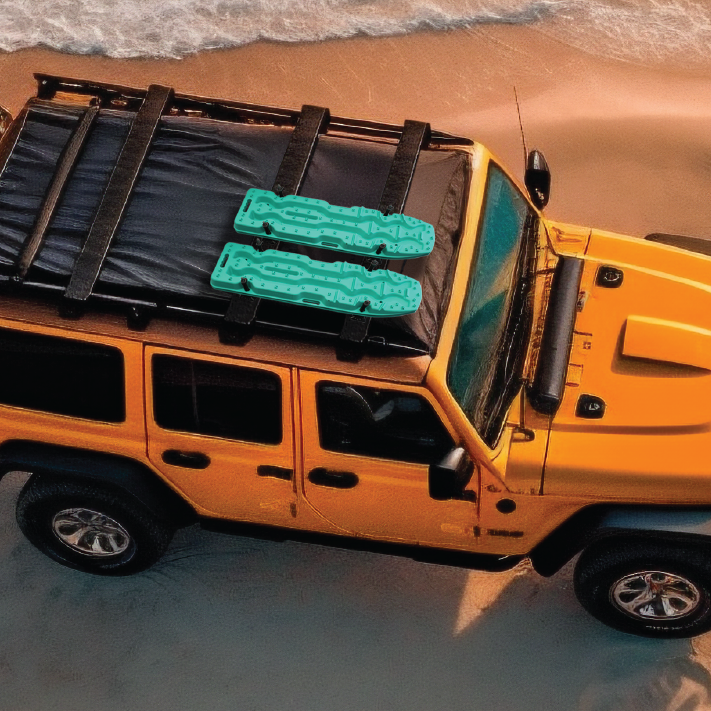 A yellow off-road vehicle parked on sand near a shoreline, featuring an Exitrax Recovery Board Ultimate 1150 - Blood Orange, Pair + Recovery Board Mounts Bundle secured to the black roof rack. Waves from the ocean are gently lapping onto the shore in the background.