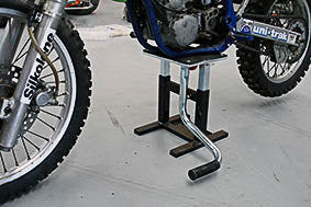 In a workshop, an Orcon Dirt Bike Lift supports a motorcycle on its platform. The close-up shot highlights the bike's rear and side, showcasing its branding and mechanical parts. The lift stand is in black and silver tones and includes a foot pedal operation with a convenient handle for ease of use.