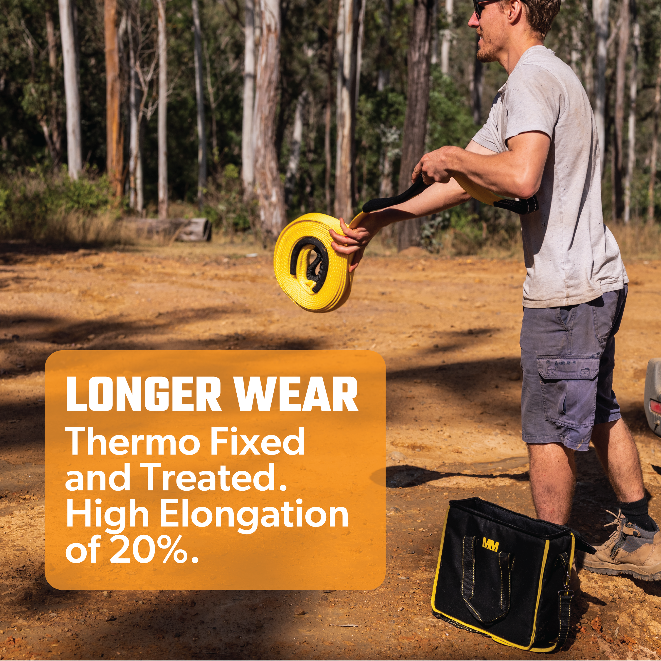 A person wearing a gray t-shirt and shorts handles the Mean Mother Snatch Strap 8,000kg in a scenic outdoor setting, surrounded by trees. A black and yellow bag rests on the ground nearby. Text overlay reads: "Longer Wear. Thermo Fixed and Treated Nylon Webbing with High Elongation of 20%.