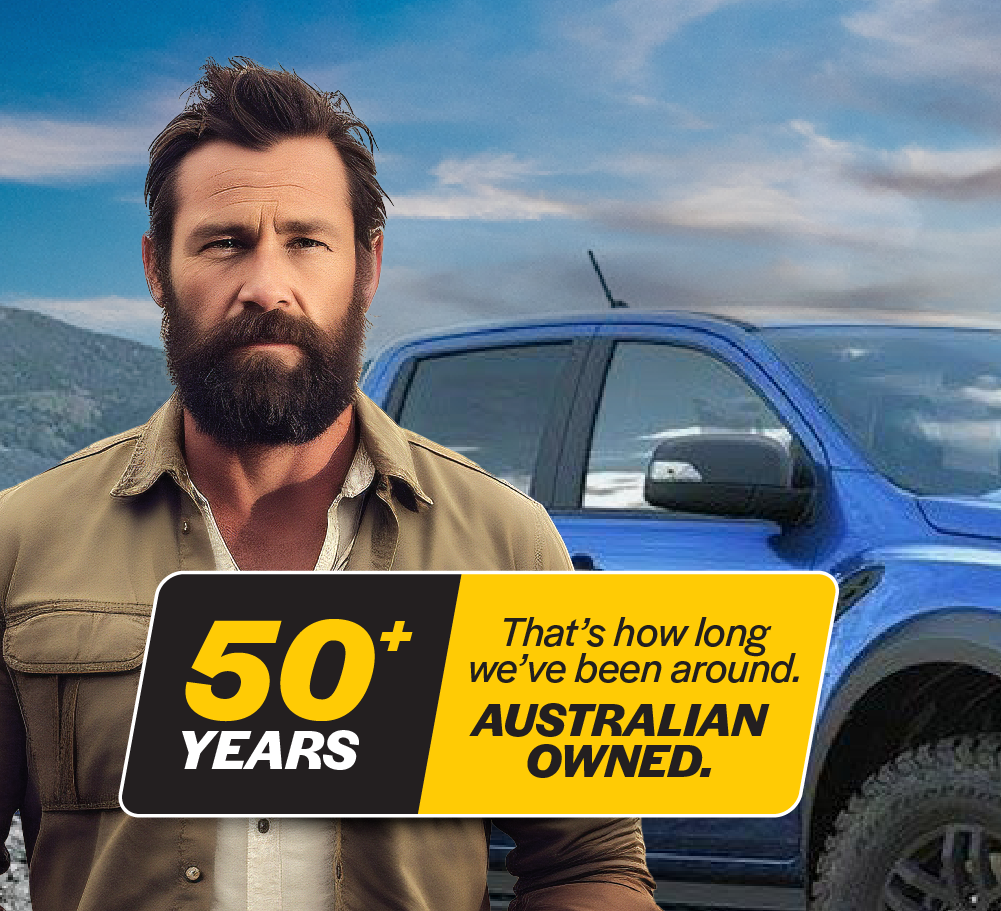 A bearded man stands in front of a blue truck with mountains in the background. A yellow and black sign beside him reads "50+ YEARS That's how long we've been around. AUSTRALIAN OWNED." The truck's equipped with premium 4x4 gear including a Mean Mother DCDC Charger 20 Amp (Trade Show Only) for rugged adventures.