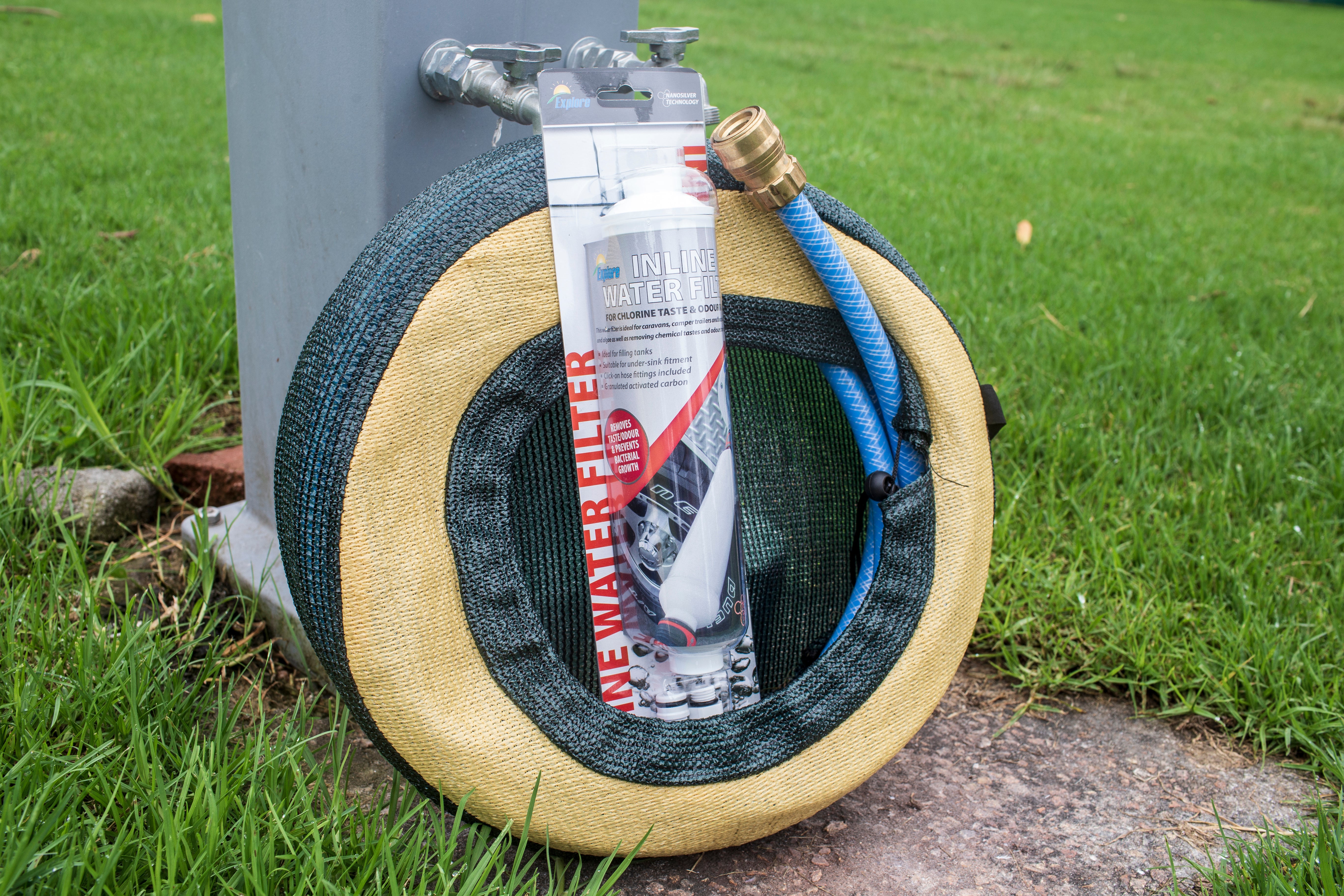 A coiled garden hose hangs on a metal post, connected to a water spigot. Attached to the hose is a package of the Explore Inline Water Filter - Nano Silver, suitable for RV or garden use. The scene is set on a patch of green grass with a concrete area underneath.