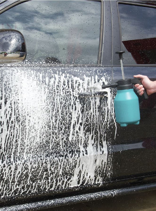 Using the Mean Mother 4x4 Pressure Foam Sprayer 2L, a person sprays thick white foam onto the side of a dark car with a blue handheld pump sprayer, prepping the vehicle for cleaning.