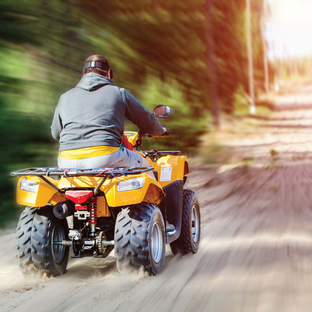 A person wearing a grey hoodie and black cap rides a yellow all-terrain vehicle (ATV) down a dirt path surrounded by trees. Attached to the ATV is the Mean Mother ATV Winch 2000lb with Synthetic Rope, ready for action. The background appears blurred, giving a sense of speed and motion, with late afternoon sunlight filtering through the foliage.