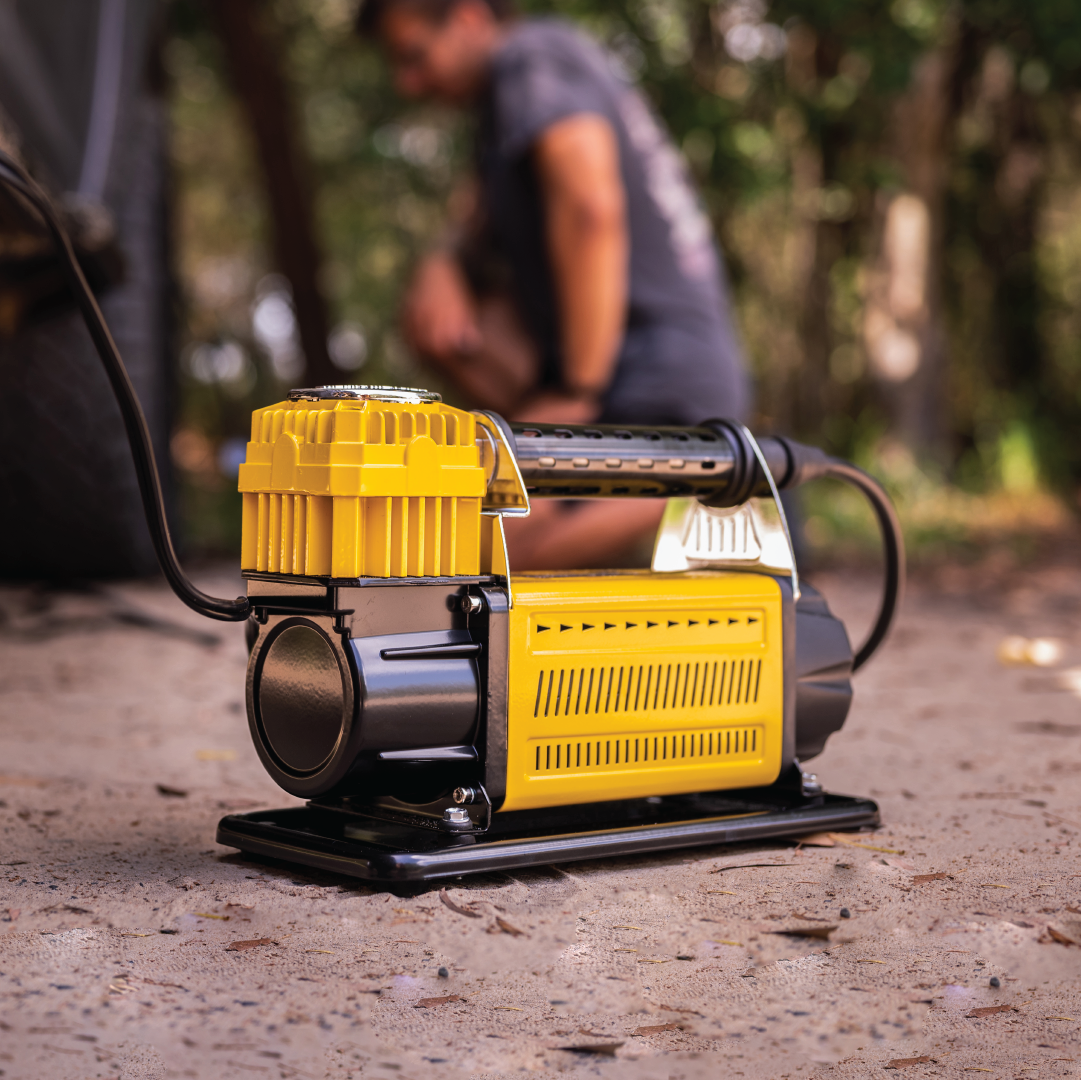 A Mean Mother Air Compressor with Wireless Remote Control, featuring a robust 180L/min free air delivery and housed in a yellow casing, is stationed on a concrete surface outdoors. In the background, an individual is seen kneeling by a vehicle tire, making adjustments. This reliable compressor comes with the added assurance of a 5-year warranty. Trees and greenery form a blurred backdrop in the distance.