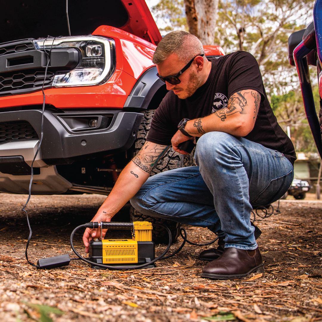 A person in a black T-shirt and jeans kneels beside a red vehicle with the hood open, using the Mean Mother Air Compressor with Wireless Remote Control 180lpm to inflate a tire. The powerful 180L/Min free air delivery ensures quick results as they work outdoors, surrounded by trees.