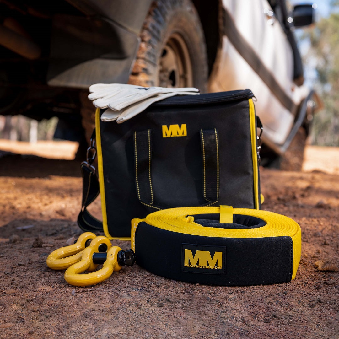 A Mean Mother 4x4 4WD Recovery Kit 8,000kg 5pc is displayed on the ground near a vehicle. It features a black and yellow bag, snatch strap, yellow shackles, and a pair of gloves. The scene unfolds on a rugged dirt path.