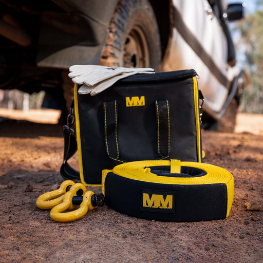 A Mean Mother 5pc Recovery Kit 8,000kg in black and yellow sits on the ground, perfect for any 4WD enthusiast. The kit includes a branded storage bag, recovery straps, a snatch strap, yellow shackles, and a pair of white gloves. In the background, a vehicle tire and part of a vehicle rest on the dirt surface.