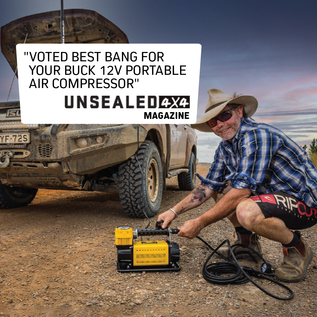 A man kneels on a dirt road beside a rugged SUV, inflating its tire using a Mean Mother Adventurer 4 – 180lpm Air Compressor Bundle. The image features a badge that reads: "Voted Best Bang for Your Buck 12V Portable Air Compressor" by Unsealed 4X4 Magazine.