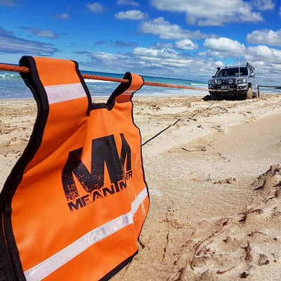 A four-wheel drive vehicle is parked on a sandy beach, linked with a snatch strap. In the foreground, an orange recovery device from the "Mean Mother 4x4" brand is visible. The partly cloudy sky creates an ideal setting for any 4WD enthusiast.